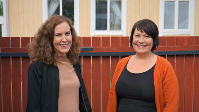 Två kvinnor står framför ett brunt staket.