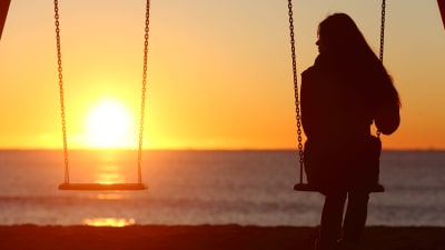 en kvinna sitter på en gunga vid stranden, det är solnedgång. Gungan bredvid henne är tom. 