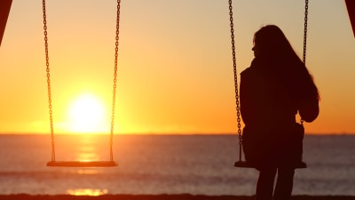 Kvinna sitter på gunga i solnedgång, bredvid henne en tom gunga