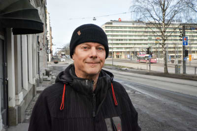 Man i svart jacka och mössa står på gata i stad och han tittar in i kameran.