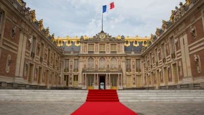 Versaillesin linnan sisäänkäynti, jonka eteen on levitetty punainen matto. Katolla liehuu Ranskan lippu.