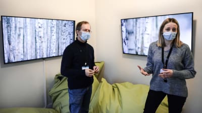 En man och en kvinna i munskydd står inne i ett rum och berättar något mot kameran. I bakgrunden syns stora gröna kuddar och två skärmar på väggen som visar naturbilder.