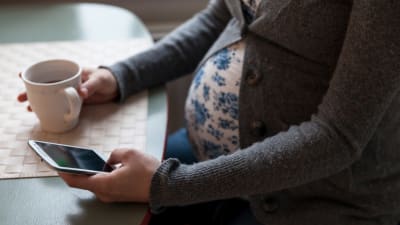 Gravid kvinna sitter och dricker kaffe och använder sin mobiltelefon.