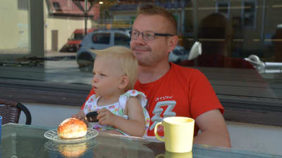 Janne Ylönen sitter vid ett bord och tittar ut mot vägen, han har sitt gudbarn i famnen.