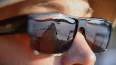 En man i solglasögo, som reflekterar en bild av en sjö och en brygga.