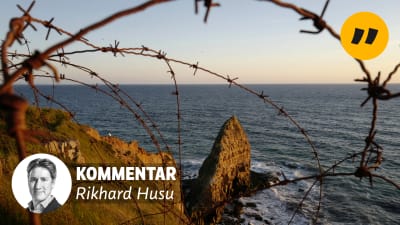 Taggtråd ligger på klippor vid stranden. På bilden en banner med texten "Kommentar Rikhard Husu".