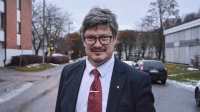 Medelålders man i kostym står på väg, tittar in i kameran med ett litet leende.