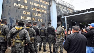 Soldater i utrustning fotade bakifrån. De står utanför en fängelsemur och väntar på att släppas in på området.  