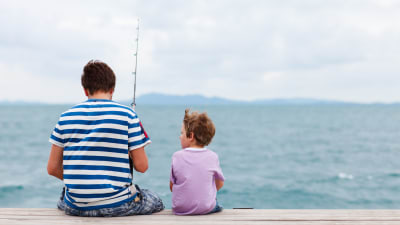 En pappa sitter med sin son på en brygga med ett fiskespö. I bakgrunden syns hav och en molnig himmel.