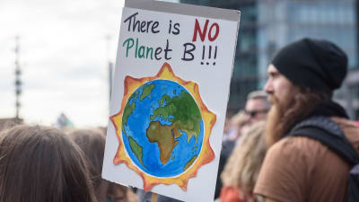 En skylt med texten "there is no planet b" (på svenska "det finns ingen planet b" och en bild på ett jordklot som brinner. Demonstranter i bakgrunden.