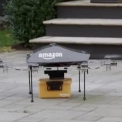 En liten drönare med Amazon.coms logotyp är på marken och har en liten orange låda nertill.
