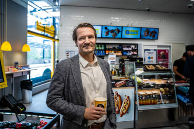 En vit medelålders man tittar mot kameran. Han håller i en kaffekopp och står inne i en R-kiosk. 