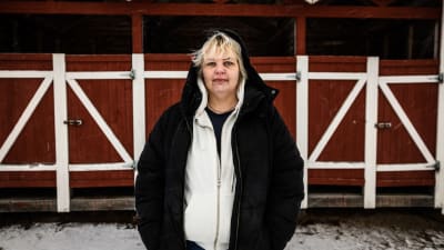 En kvinna med huva står framför ett rödvitt hus.
