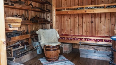 Inuti en liten stuga i trä: våningssäng, hyllor med verktyg, skinnfällbeklädd stol gjord av en gammal tunna.