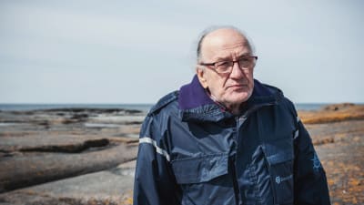 Äldre man i glasögon och blå överrock står på klippa med hav i bakgrunden.