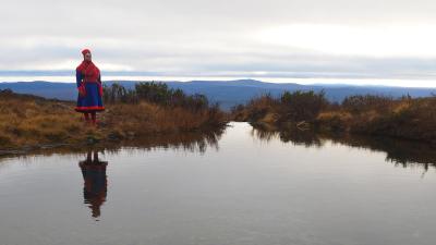 Saamenpukuinen nainen (ohjaaja Suvi West) seisoo joen rannalla syksyisessä Lapin maisemassa, taustalla sinertävät tunturit.