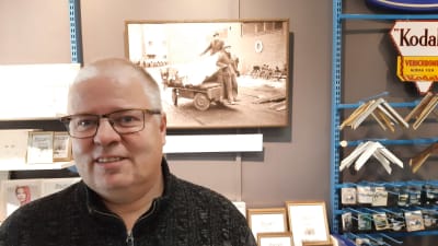 En man med kort vitt hår och glasögon tittar rakt in i kameran. I bakgrunden finns ett svartvitt foto och fotoramar på väggen.