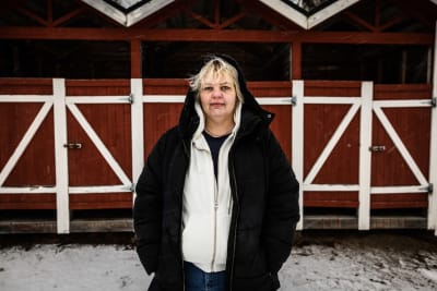 En kvinna med huva står framför ett rödvitt hus.