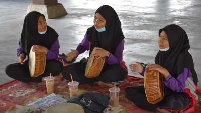 Tre kvinnor i burka sitter på en matta på golvet och spelar handtrumma.
