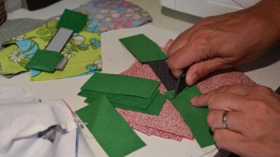 Tygbitar i grönt och rött ligger på sybordet. Den ena handen håller i en grön tygbit och den andra handen klipper av en sytråd.
