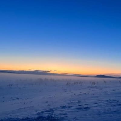 Luminen tunturimaisema keskellä kaamosta. Taivaanrannalla näkyy oranssinkeltaista valoa.