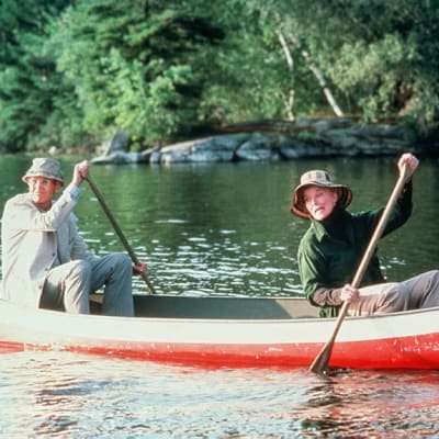 Iäkäs nainen ja mies melovat punavalkoisella kanootilla tyynellä järvellä, naisen istuessa edessä ja sanoen jotain miehelle.
