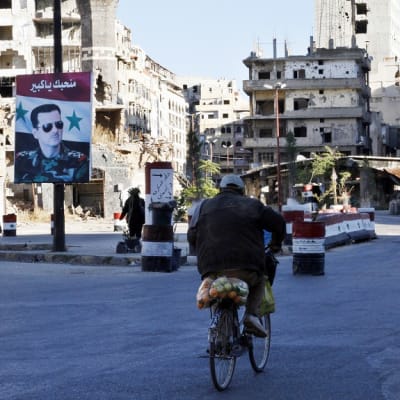 Kuva Homsin vanhasta kaupungista. Kuvan keskellä lippalakkipäinen mies ajaa polkupyörällä kohti hallituksen tarkastuspistettä, jossa on ainakin yksi univormupukuinen mies. Polkupyörän tarakalla on hedelmiä läpinäkyvässä muovipussissa. Myös pyörän sarvista roikkuu pussi. Pylväässä tarkastuspisteen kohdalla on suuri juliste Syyrian presidentistä Bashar al-Assadista sotilasasussa mustat lasit päässään. Tarkastuspistettä reunustavat tynnyrit on maalattu Syyrian lipun väreihin. Taustalla näkyvät rakennukset ovat tulituksen jäljiltä täynnä reikiä. Aurinko paistaa mutta varjot ovat pitkiä.