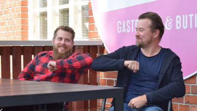 Köttkontrollens restaurangchef Mikael Kokkonen gör den såkallade coronahälsningen med affäremannen Olli Muurainen på Köttkontrollens terass. Två män rör vid varandras armbåge, sittande framför en tegelvägg.