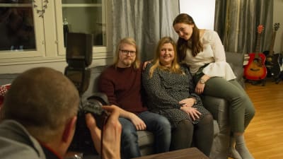 En kvinna sitter med sina två vuxna barn i en soffa. En fotograf tar bild på dem.
