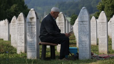 En man med muslimsk mössa på huvudet sitter på en pall och läser en bok brevid vita, obeliskformade gravstenar.