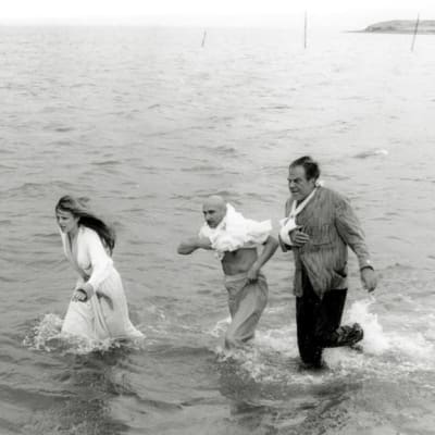 Nuori nainen ja kaksi keski-ikäistä miestä juoksevat polviin ulottuvassa vedessä meren rannalla.