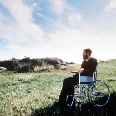Parrakas mies tummissa vaatteissa istuu pyörätuolissa katsellen kallioista ja ruohikkoista nummimaisemaa.