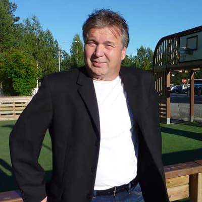 Mustaan pukutakkiin pukeutunut mies nojaa urheilualueen puiseen aitaan kesällä.