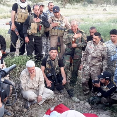 Joukko sotilaita seisoo ruohikkoisessa maastossa haudan edessä päät painuksissa, monet pitävät toista kättään rinnallaan kunnioituksen osoituksena. Osalla miehistä on maastopuvut, osalla siviilivaatteet, osalla mustat puvut. Haudalla on ilmeisesti Irakin lippu.