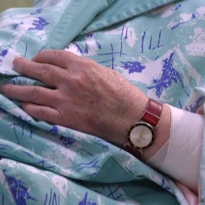 Sairaalassa olevan vanhuksen käsi, jossa on kello ja side.