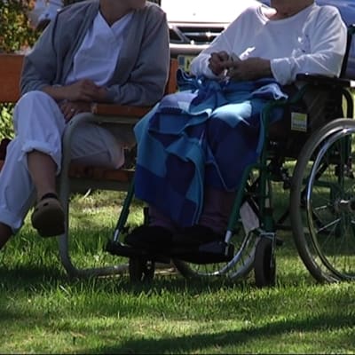 Sairaanhoitajan valkoiseen asuun pukeutunut hoitaja istuu puistossa pyörätuolissa istuvan vanhuksen kanssa.