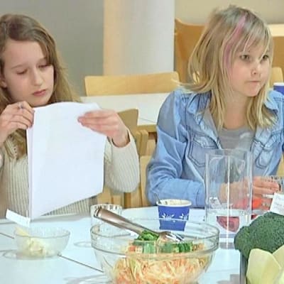Kaksi tyttöä istuu koulun ruokalassa, toinen katsoo paperia.