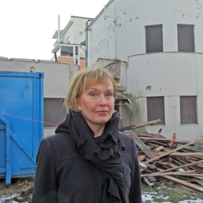 Kuvassa nainen lautakasan ja ränsistyneen rakennuksen edessä