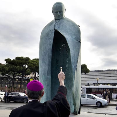Pappi siunaa vastapaljastetun paavi Johannes Paavali II:sta esittävän patsaan Rooman päärautatieaseman edessä.
