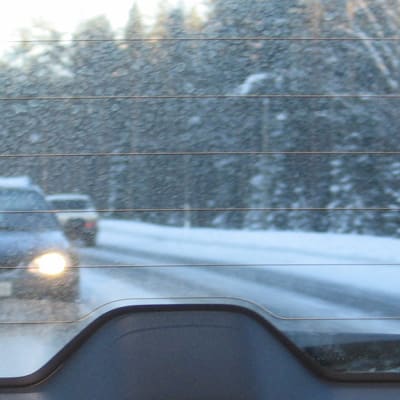 Kuvassa auton takalasin läpi kuvattu takaa tuleva auto. Lumi pöllyää. 