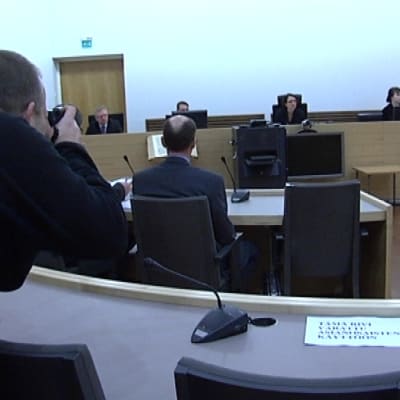Valokuvaaja kurkottaa ottamaan kuvaa oikeussalissa.
