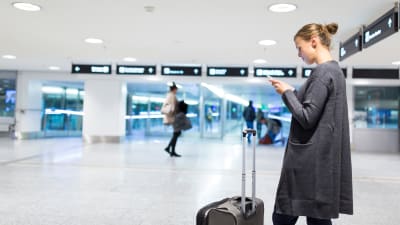 En kvinna står på ett flygfält och tittar på sin mobil, med kappsäcken framför sig.