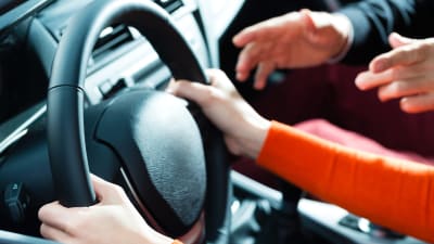 En ung kvinnas händer vid ratten på en bil med en äldre mans händer som gestikulerar från passagerarsätet. 