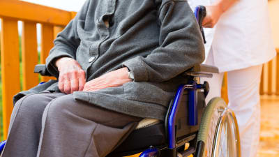 Äldre person sitter i en rullstol som en vårdare skuffar fram.