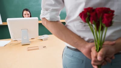 man som döljer rosor bakom sin rygg och tittar på en ung kvinna som jobbar vid sin dator