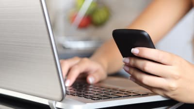 Kvinna som sitter framför en bärbar dator med en smarttelefon i handen.