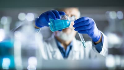 En manlig forskare med skägg och blåa handskar  häller blå vätska i ett provrör i ett laboratorium. Ansiktet syns inte. Snygg bildsättningsbild.