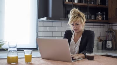 Förbryllad kvinna sitter i köket och läser något på sin dator
