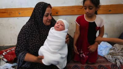 En äldre kvinna håller ett nyfött barn i famnen. Bredvid henne står ett barn. De har blivit hemlösa på grund av Israels flygräder.