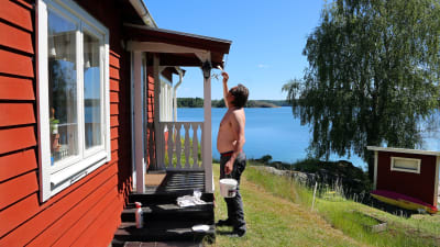 Person målar röd stuga med vita knutar vid strand. 
